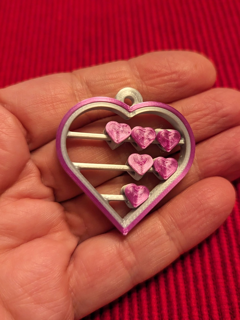 Ett hjärtformat ornamenthänge med rosa/lila hjärtan inuti en vit och lila yttre ram. Föremålet ser handgjort eller 3D-printat ut och kan fungera som en dekorativ accessoar.