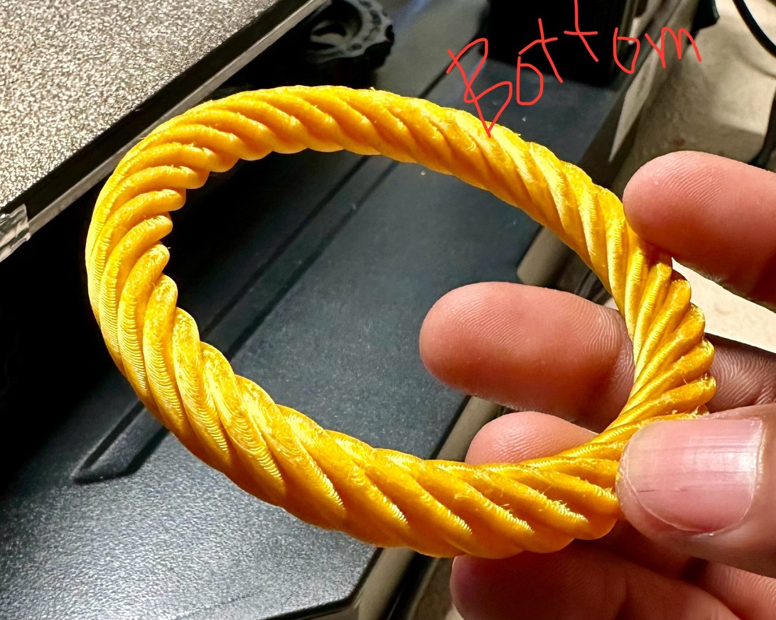 Ett ljust gult eller guldfärgat tvinnat rep-liknande armband eller armring som hålls mellan fingrarna. Föremålet har en spiralformad eller tvinnad textur och ser ut som en samtida modeaccessoar, möjligen 3D-printad eller gjord av tyg.