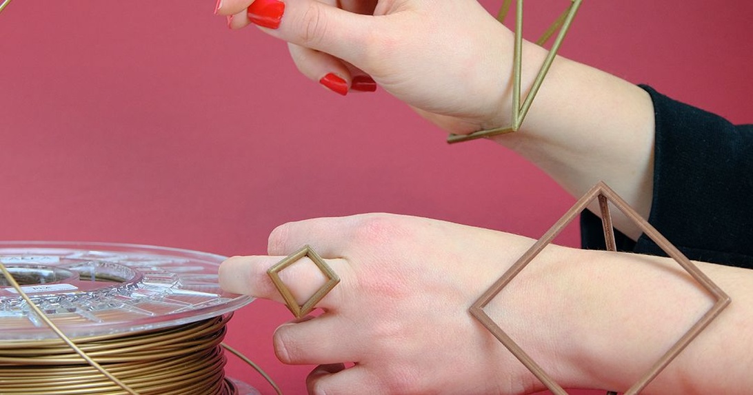 En närbild på händer med rött nagellack som arbetar med geometriska smycken i guld/mässing. Bilden visar fyrkantiga ringar och metalltråd, vilket ser ut som moderna handgjorda smycken under tillverkning.